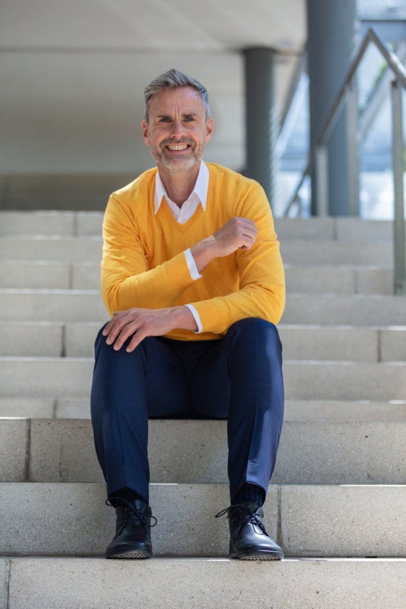 Ein Mann mit grauem Haar und Bart sitzt lächelnd auf einer Außentreppe. Er trägt einen leuchtend gelben Pullover über einem weißen Hemd und dunkelblauen Hosen. Er hat schwarze Schuhe an und sitzt lässig da, ein Arm ruht auf seinem Knie. Im Hintergrund sind eine Betonkonstruktion und Geländer zu sehen.