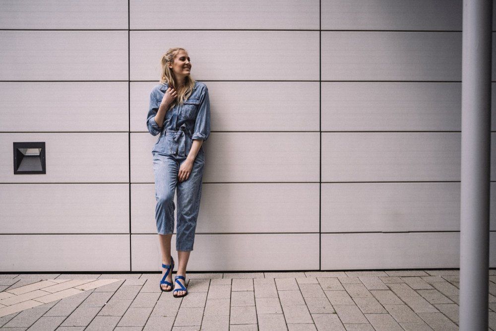 Eine Person im Jeans-Outfit und MIQA Blue Sandalen von ZAQQ lehnt an einer gefliesten Wand, eine Hand im Nacken. Sie wirkt entspannt und blickt zur Seite, während sie auf einem gepflasterten Gehweg steht.